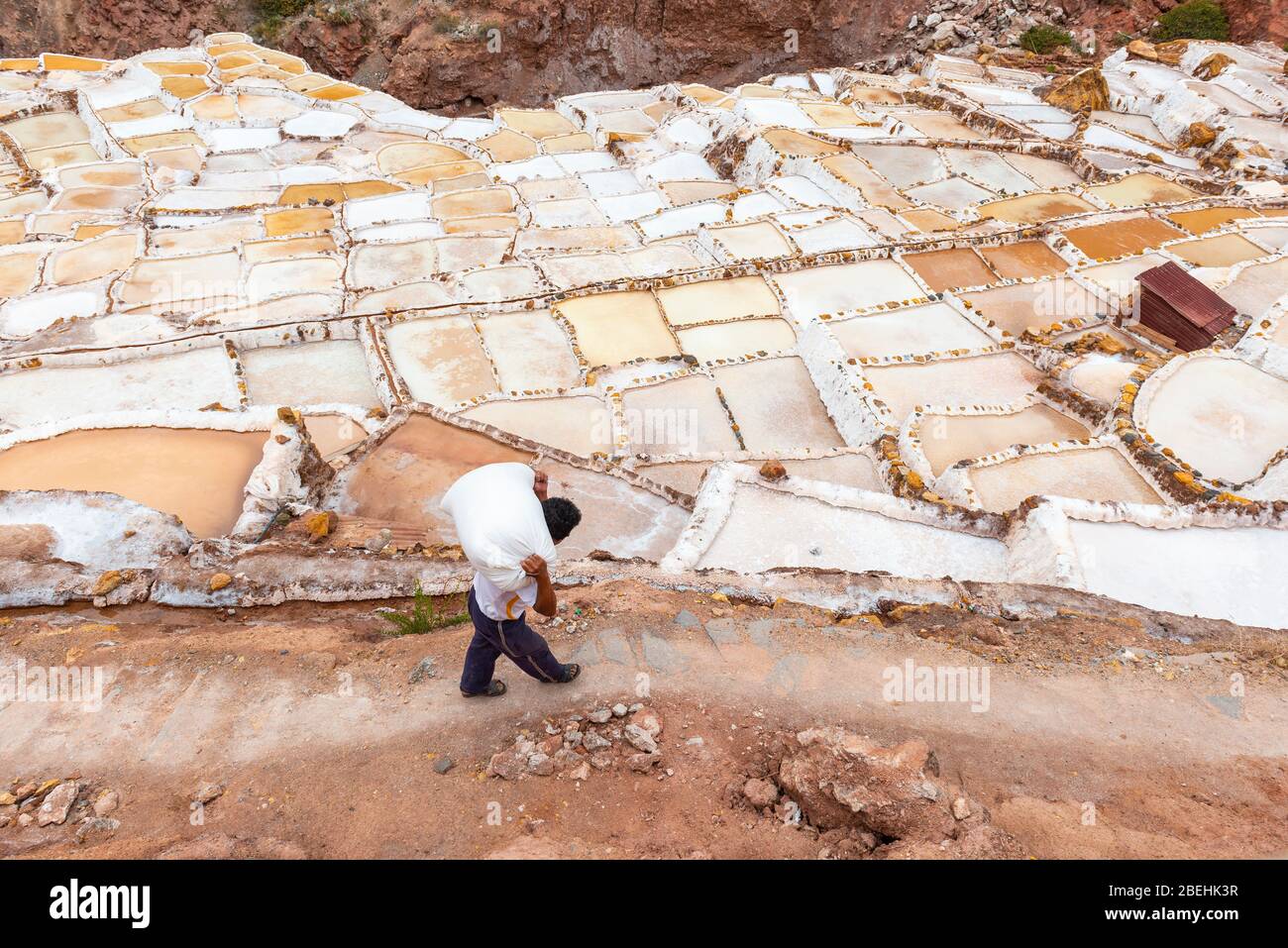 Paesaggio delle terrazze di sale di Maras con un uomo indigeno Quechua che trasporta un sacco pesante di sale, Cusco, Perù. Foto Stock