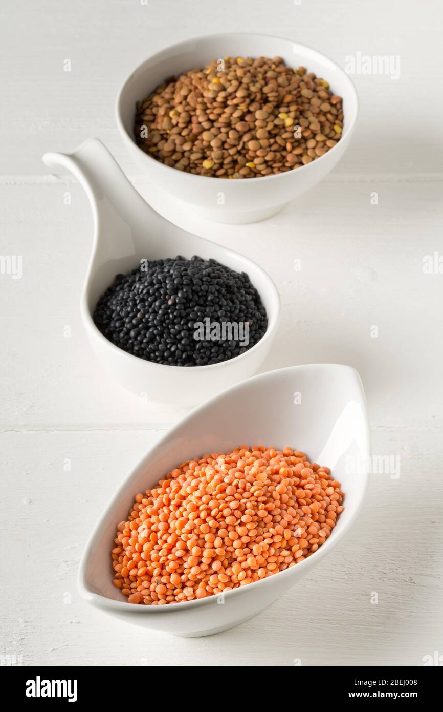 Le lenticchie assortite si mescolano con lenticchie di beluga rosse, marroni e nere in ciotole bianche su sfondo bianco tavolo di legno - fuoco selettivo Foto Stock