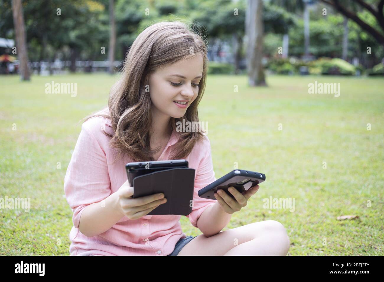 Giovane donna caucasica con smartphone in una mano e tablet in un'altra seduta a terra, sorridente. Ritratto esterno. Foto Stock
