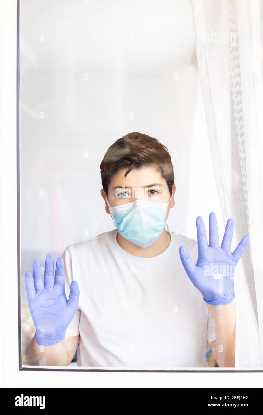 ragazzo con maschera protettiva e guanti blu che guardano fuori dalla finestra durante la quarantena del coronavirus Foto Stock