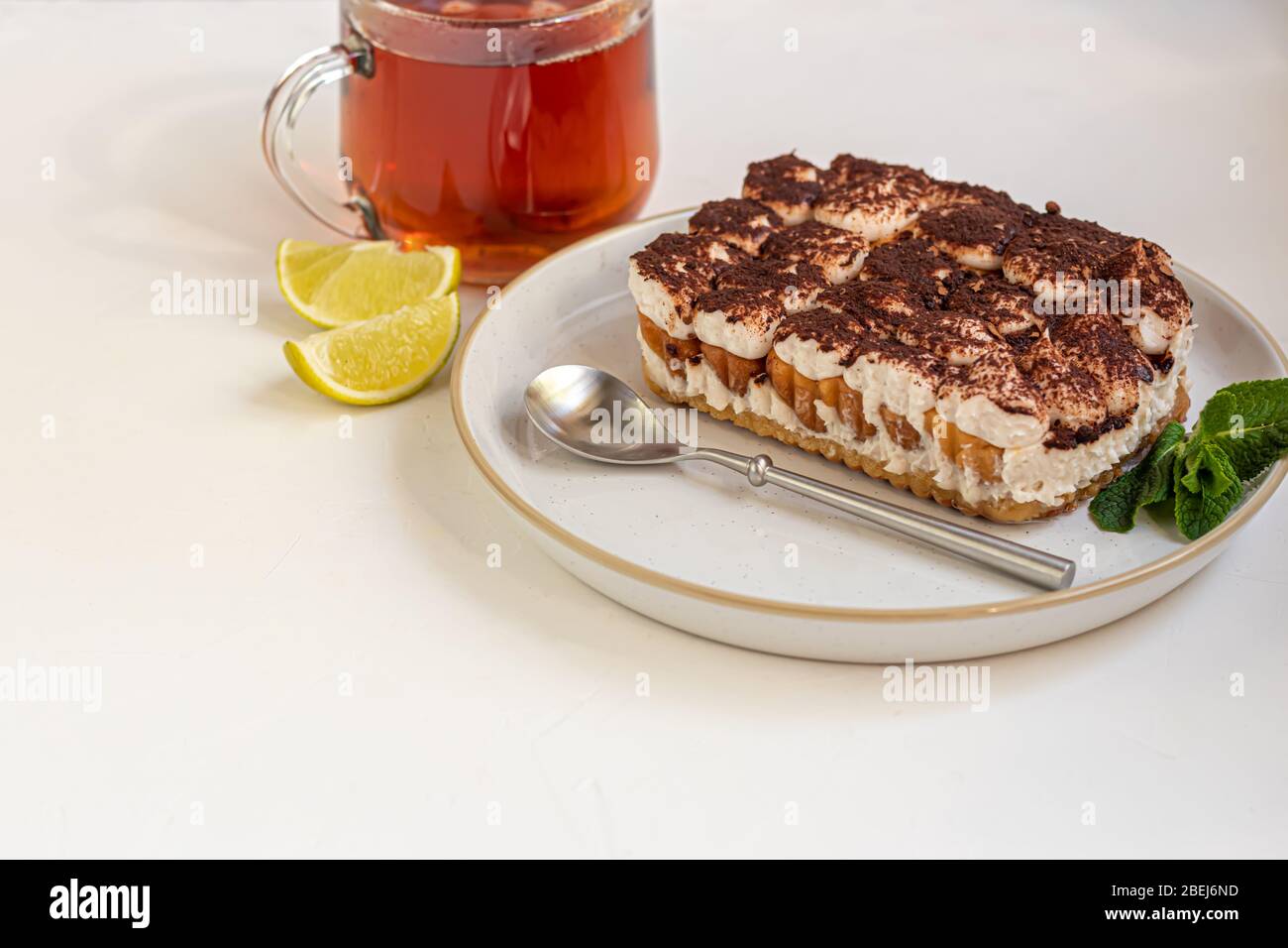 Tiramisù. Tradizionale dessert italiano su fondo bianco sotto cemento. Guarnito con un rametto di menta e fette di lime. In una tazza trasparente di tè. Foto Stock