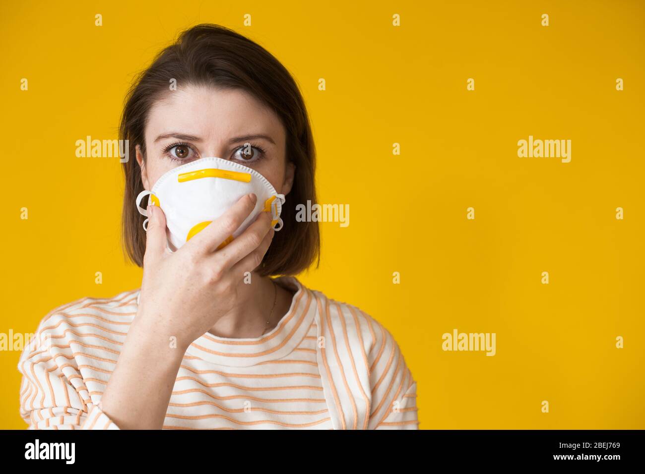 Donna caucasica con capelli neri che si posa su una parete gialla dello studio con spazio libero che tiene una maschera medica sul suo viso Foto Stock