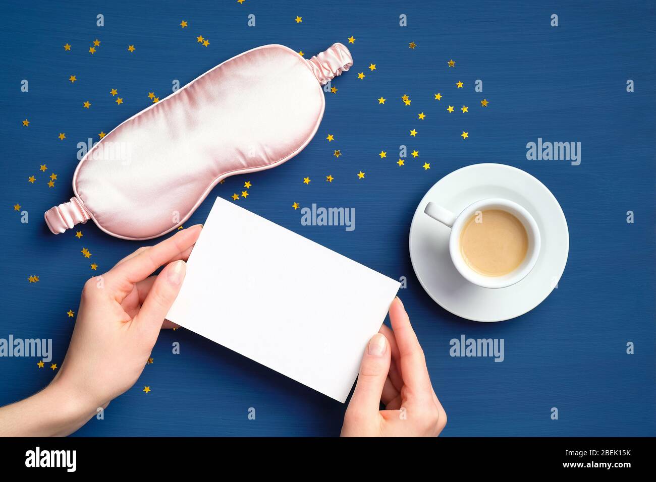 Concetto di Buongiorno. Tazza di caffè, maschera per il sonno e mani femminili che tengono carta vuota mockup su sfondo blu scuro. Disposizione piatta, vista dall'alto, copia Foto Stock