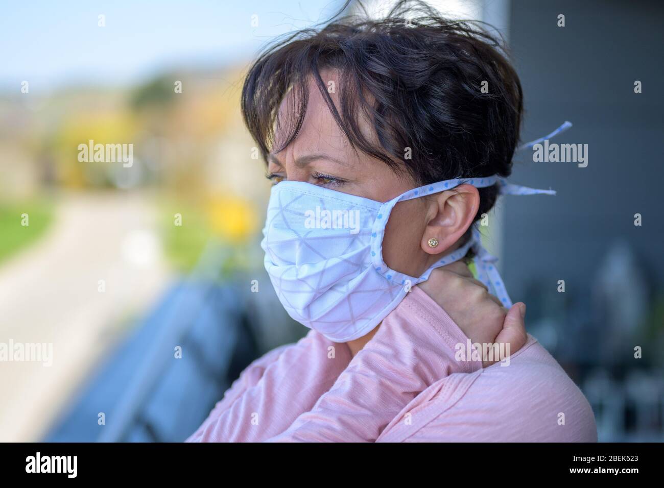 Donna ispanica che indossa una maschera chirurgica del viso, mentre si alza guardando fuori da un balcone al piano superiore come protezione contro la diffusione del coronavirus o. Foto Stock