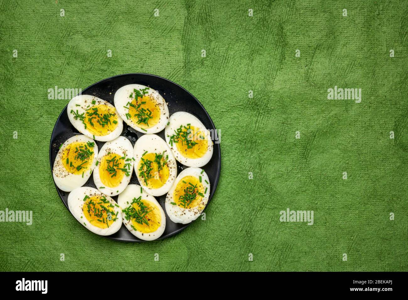 Uova di pollo bollite con erba cipollina fresca verde, sale e pepe su un piatto nero contro carta testurizzata fatta a mano - Pasqua o primavera concetto Foto Stock