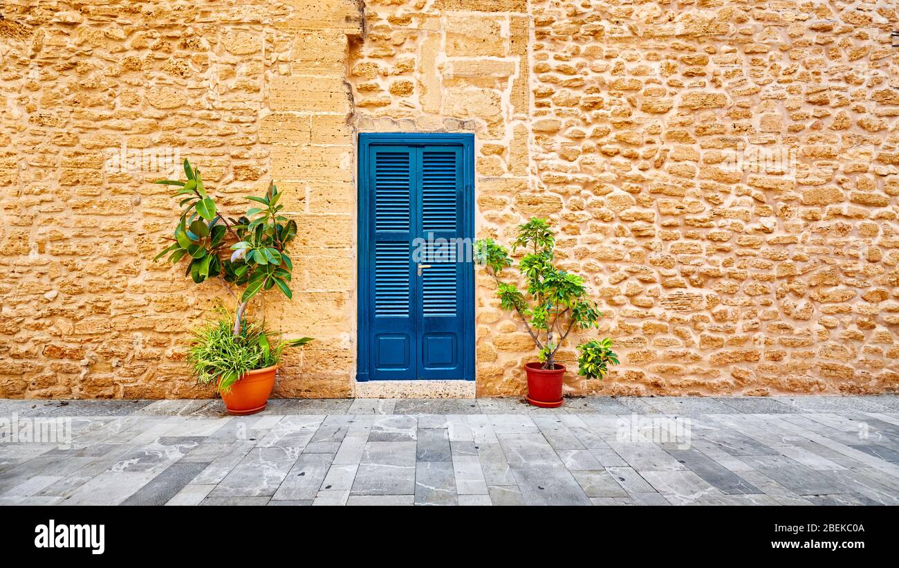 Antico muro di pietra con porta blu e fiori di vaso in una strada nel centro storico di Alcudia, Maiorca, Spagna. Foto Stock