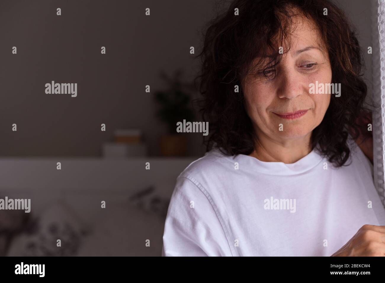 Una donna adulta triste sta in piedi accanto alla finestra nella stanza. Una persona mostra emozioni tristi. Spazio libero per il testo. Messa a fuoco morbida . Foto Stock