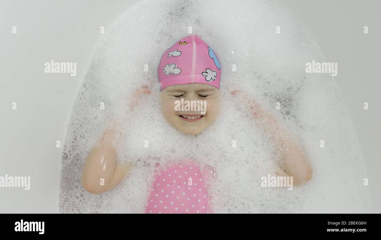 Attraente ragazza di quattro anni prende un bagno con schiuma bagno in swimwear rosa e cuffia da bagno. Sdraiati nel bagno. Igiene per il bambino biondo carino. Ragazza carina sorridente. Piccola bambina bionda in bagno Foto Stock