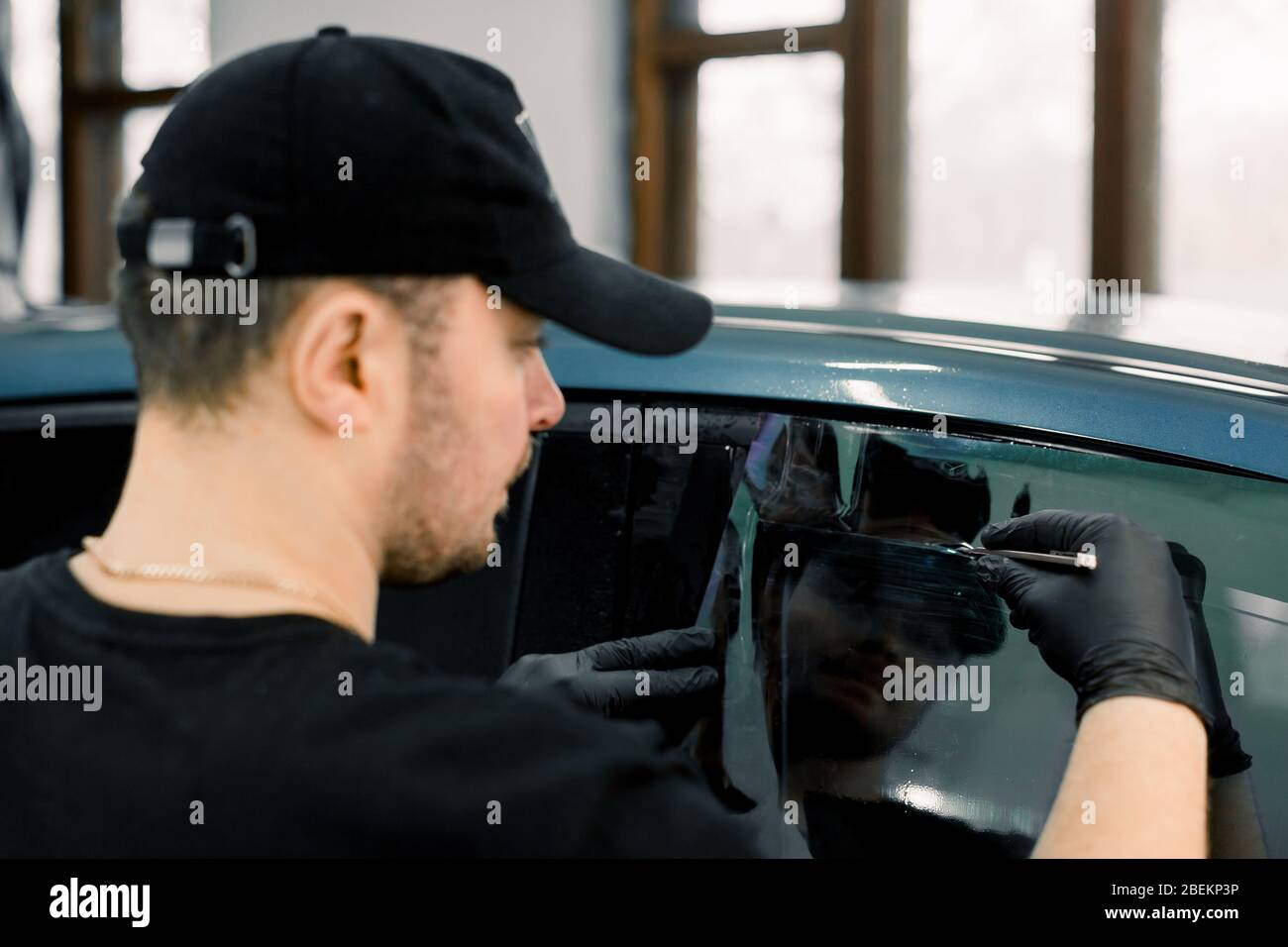 Vista posteriore dell'angolo dell'automobile che descrive l'officina del lavoratore maschio, che indossa il cappuccio nero e la t-shirt, applicando la pellicola di tinta su un finestrino dell'automobile in un garage Foto Stock