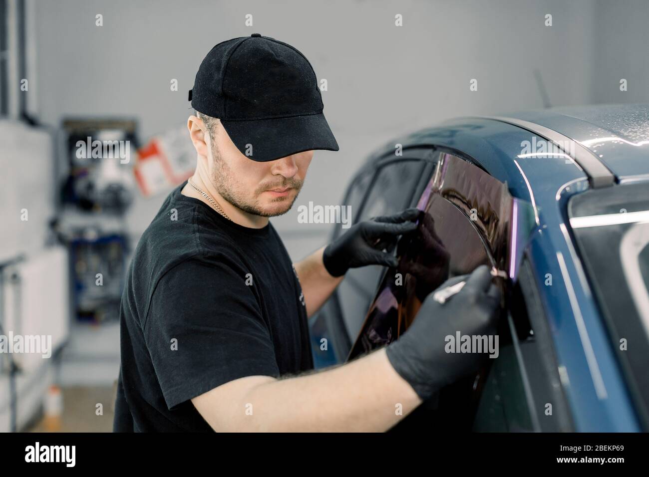Immagine di primo piano di un bel lavoratore meccanico auto, indossare uniforme nera, fissare la pellicola di tinta al finestrino auto in stazione di servizio specializzata Foto Stock