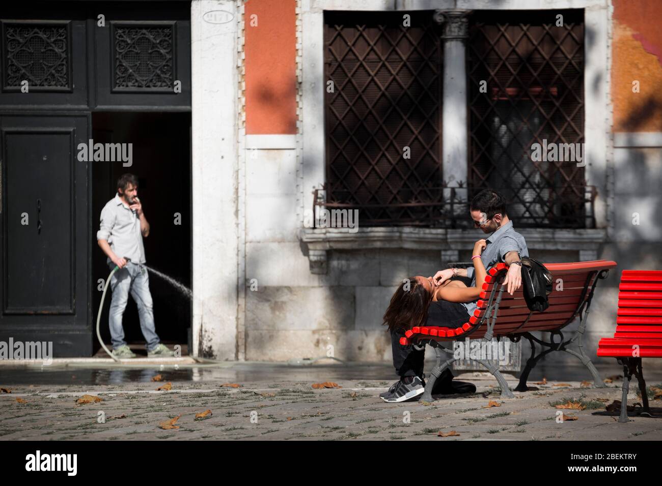 Amanti su una panchina rossa con un uomo che tiene un tubo guardando, Venezia, Italia Foto Stock