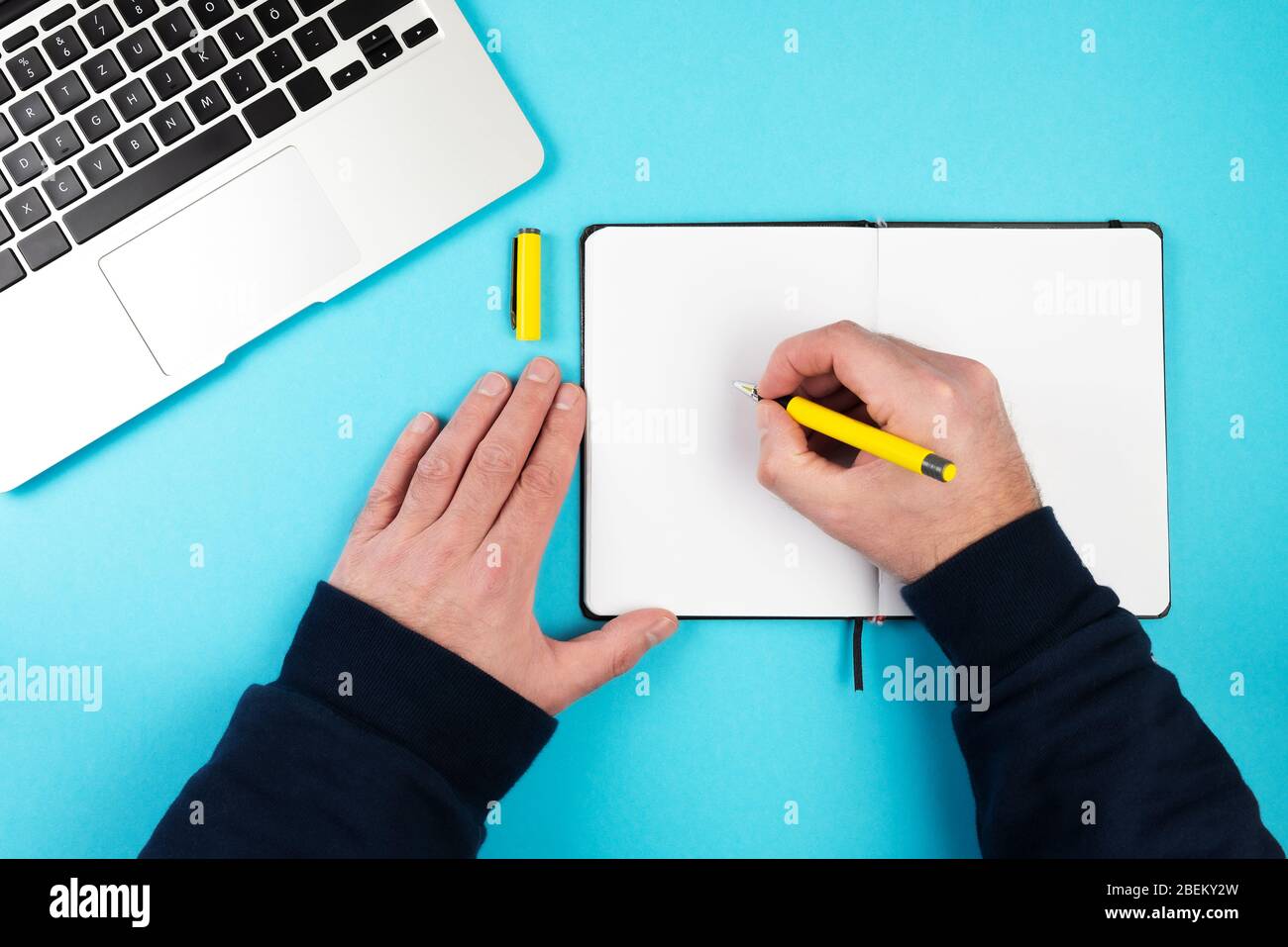 direttamente sopra lo scatto della persona che prende appunti con la penna e la carta sulla scrivania blu con computer portatile Foto Stock