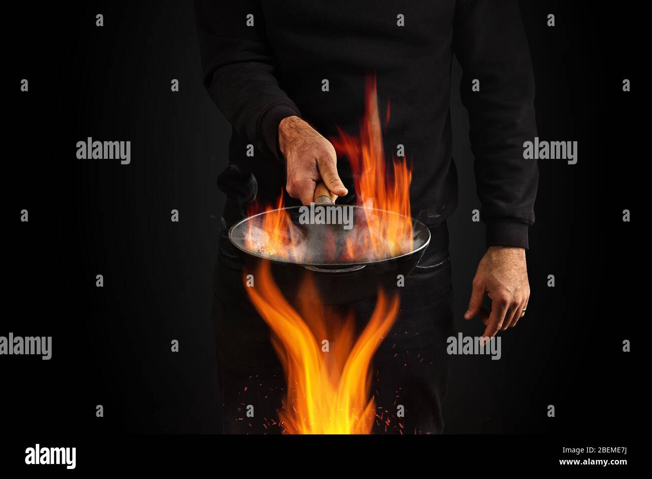 Giovane uomo vestito con jumper e jeans. Sta tenendo il wok pan bruciante con fumo sopra il fuoco, contro sfondo nero studio. Concetto di cucina. Primo piano Foto Stock