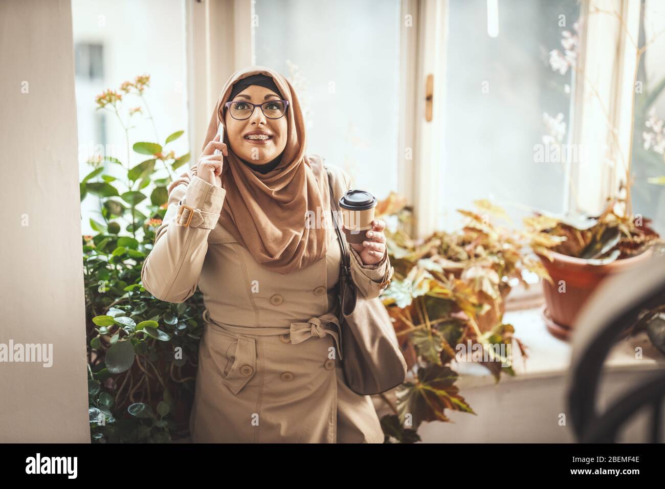 Donna musulmana di mezza età che indossa hijab con un volto felice è in piedi dalla finestra che prende il suo smartphone. Foto Stock