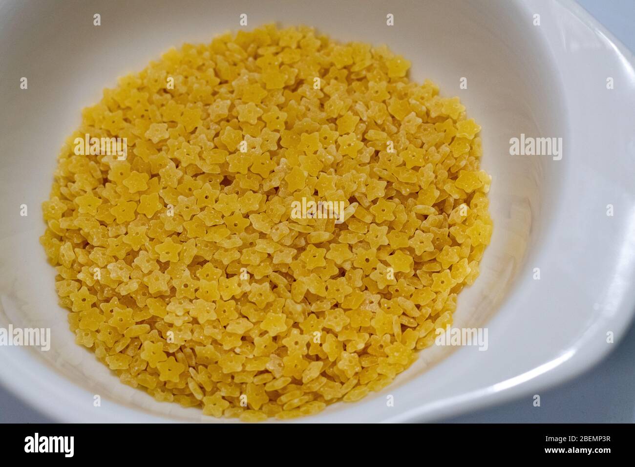 Un piccolo piatto bianco in ceramica contiene pasta stelline secca e cruda. Questa variante più piccola della pasta di zuppa di stelle ha la forma di piccole stelle. Foto Stock