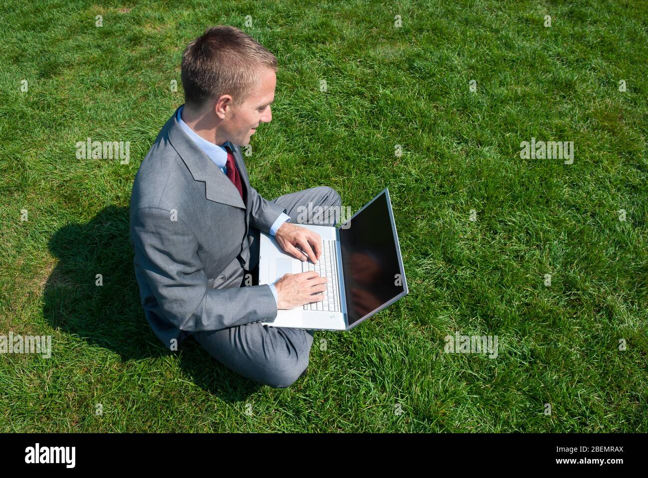 Vista a spalla di un uomo d'affari seduto all'aperto in una zona soleggiata di verde che batte sul suo computer portatile Foto Stock