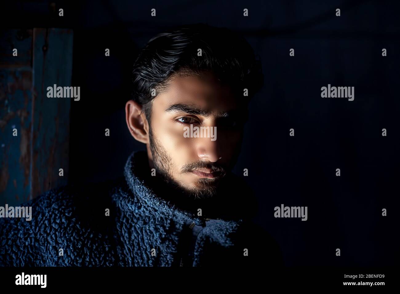 Ritratto di giovane barba maschile modello indossando un maglione di lana blu scuro e posando girato con tecnica di luce Rembrandt sul suo viso in nero Foto Stock