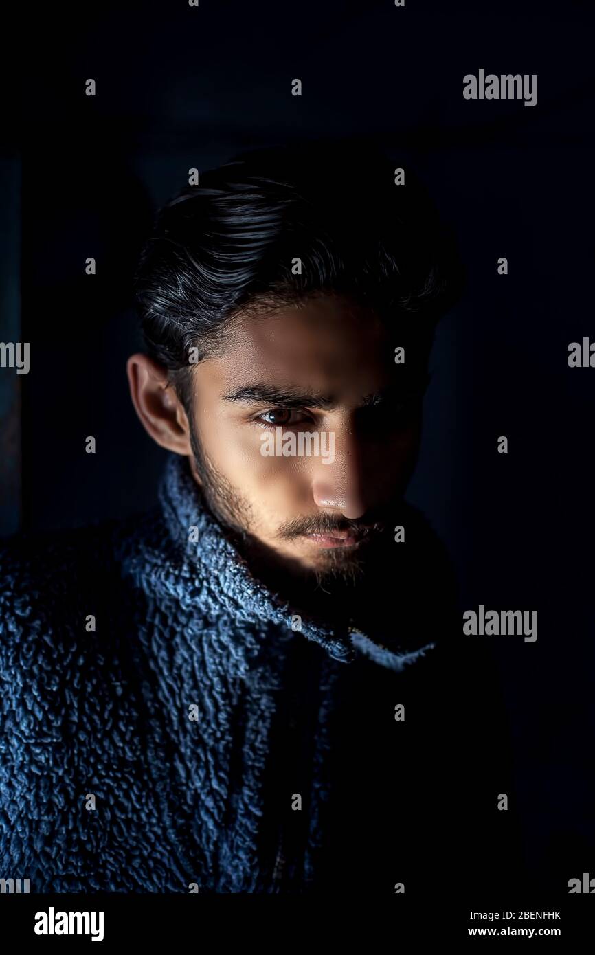 Ritratto di giovane barba maschile modello indossando un maglione di lana blu scuro e posando girato con tecnica di luce Rembrandt sul suo viso in nero Foto Stock