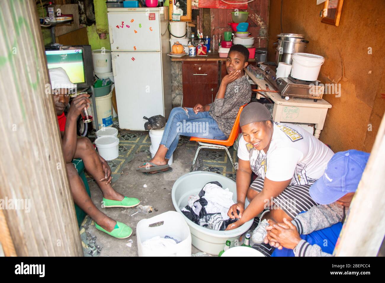 Una famiglia rilassarsi nella loro camera singola casa in una cittadina a Città del Capo. La sistemazione è un sacco con tutto il necessario presente. Le condizioni sono squallidi, ma c'è un'accettazione e i residenti sorridono. Una madre lava il bucato in una ciotola sul pavimento. Foto Stock