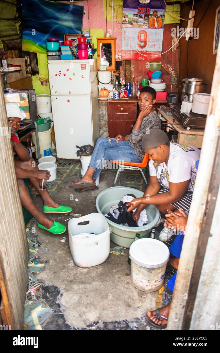 Una famiglia rilassarsi nella loro camera singola casa in una cittadina a Città del Capo. La sistemazione è un sacco con tutto il necessario presente. Le condizioni sono squallidi, ma c'è un'accettazione e i residenti sorridono. Una madre lava il bucato in una ciotola sul pavimento. Foto Stock