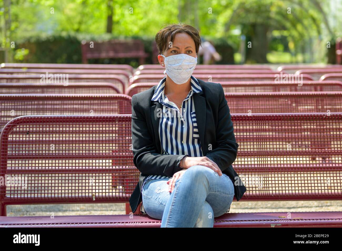 Donna elegante che indossa una maschera facciale in un parco mentre si rilassa in un luogo all'aperto con file di panche vuote durante il coronavirus o Covid-19 pandemic Foto Stock