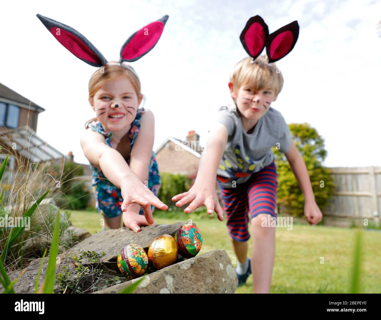Un fratello e una sorella, indossando la vernice del viso e le orecchie conigliate fatte in casa, prendere parte a una caccia all'uovo di Pasqua nel loro giardino. Foto Stock