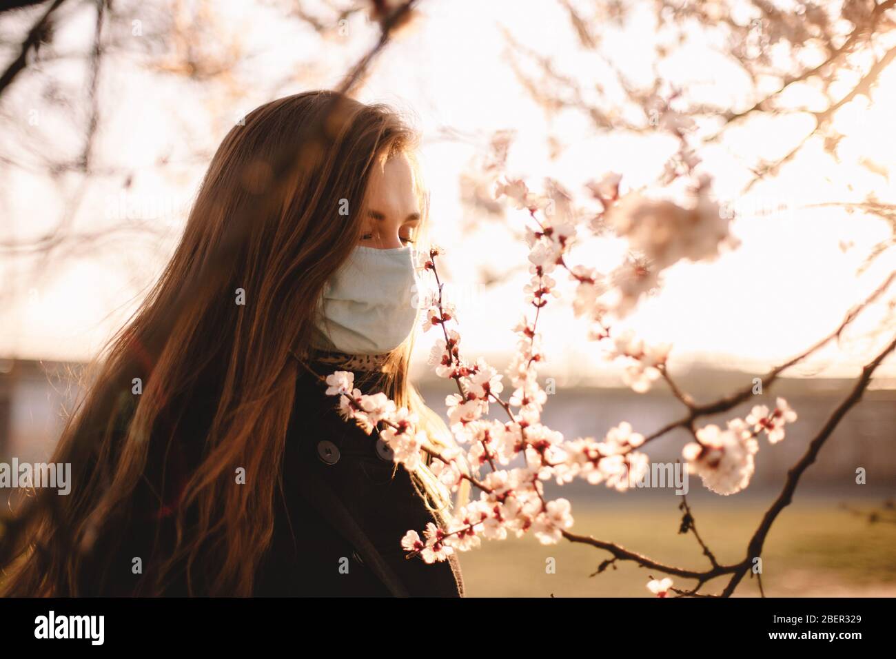 Giovane donna triste indossare viso maschera medica che odorano fiori durante la primavera Foto Stock