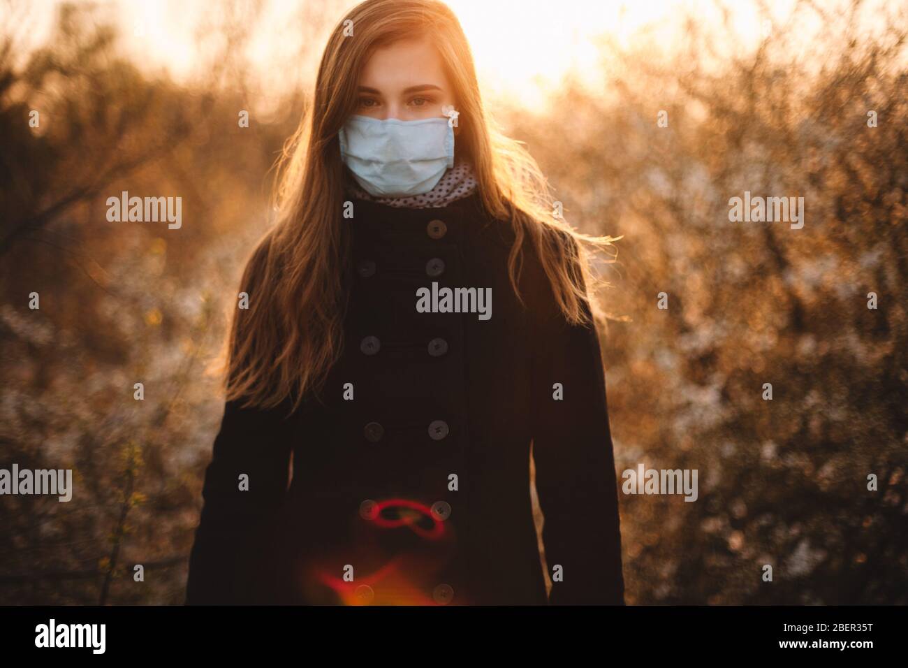 Ritratto di donna giovane triste che indossa maschera protettiva viso medico in piedi nel parco al tramonto durante la primavera Foto Stock