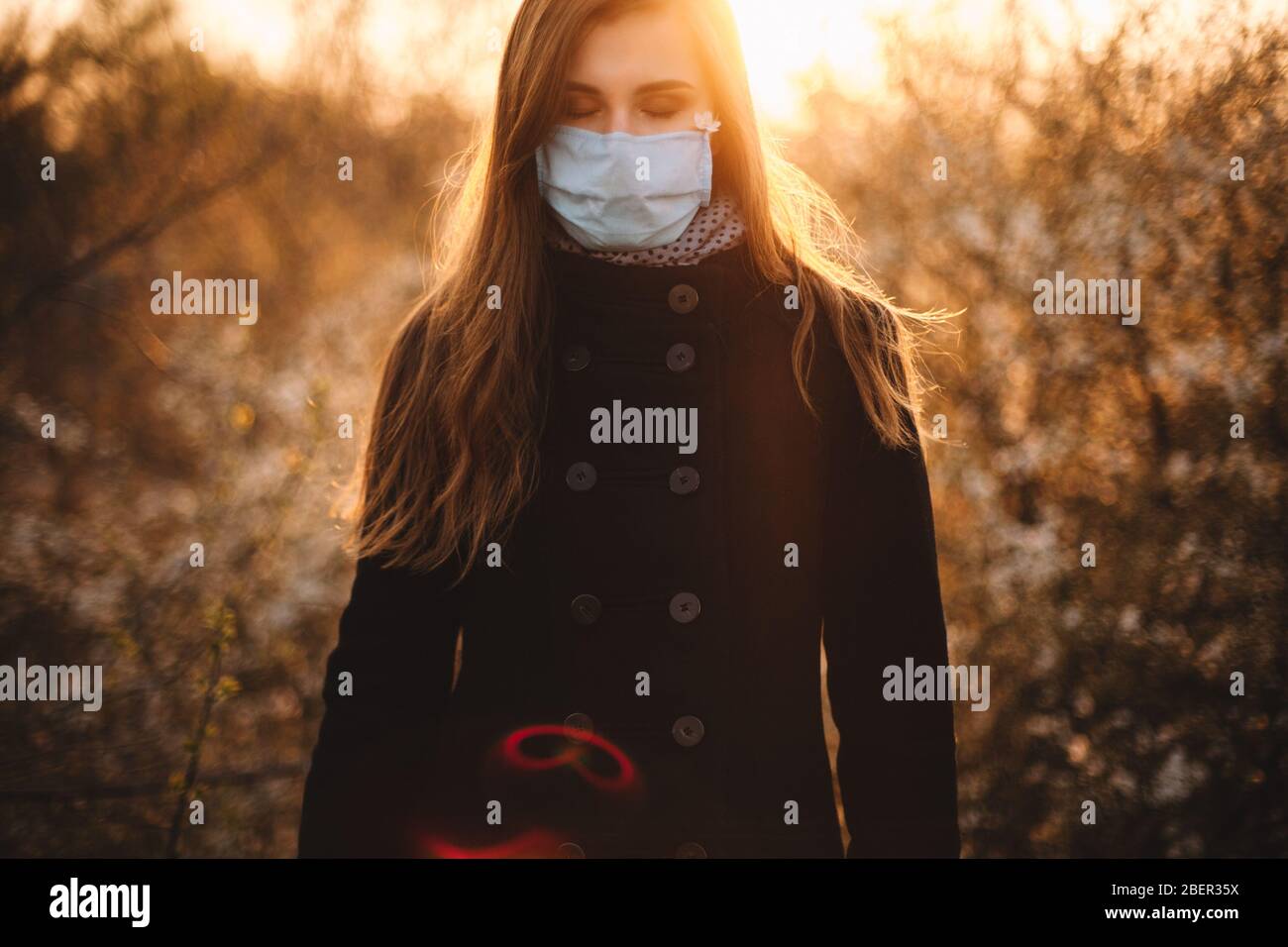 Ritratto di donna giovane triste che indossa maschera protettiva viso medico in piedi nel parco al tramonto durante la primavera Foto Stock