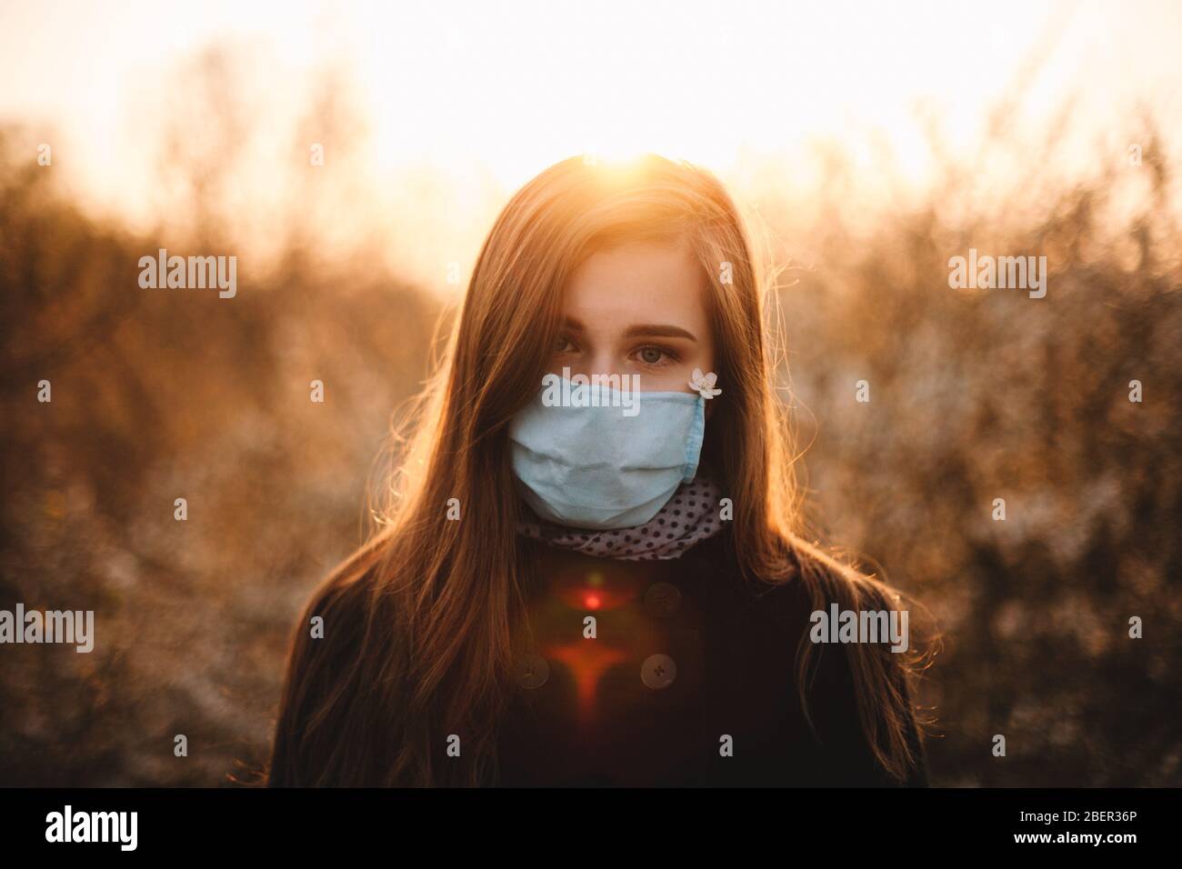 Ritratto di donna giovane cute che indossa maschera protettiva viso medico in piedi nel parco al tramonto durante la primavera Foto Stock