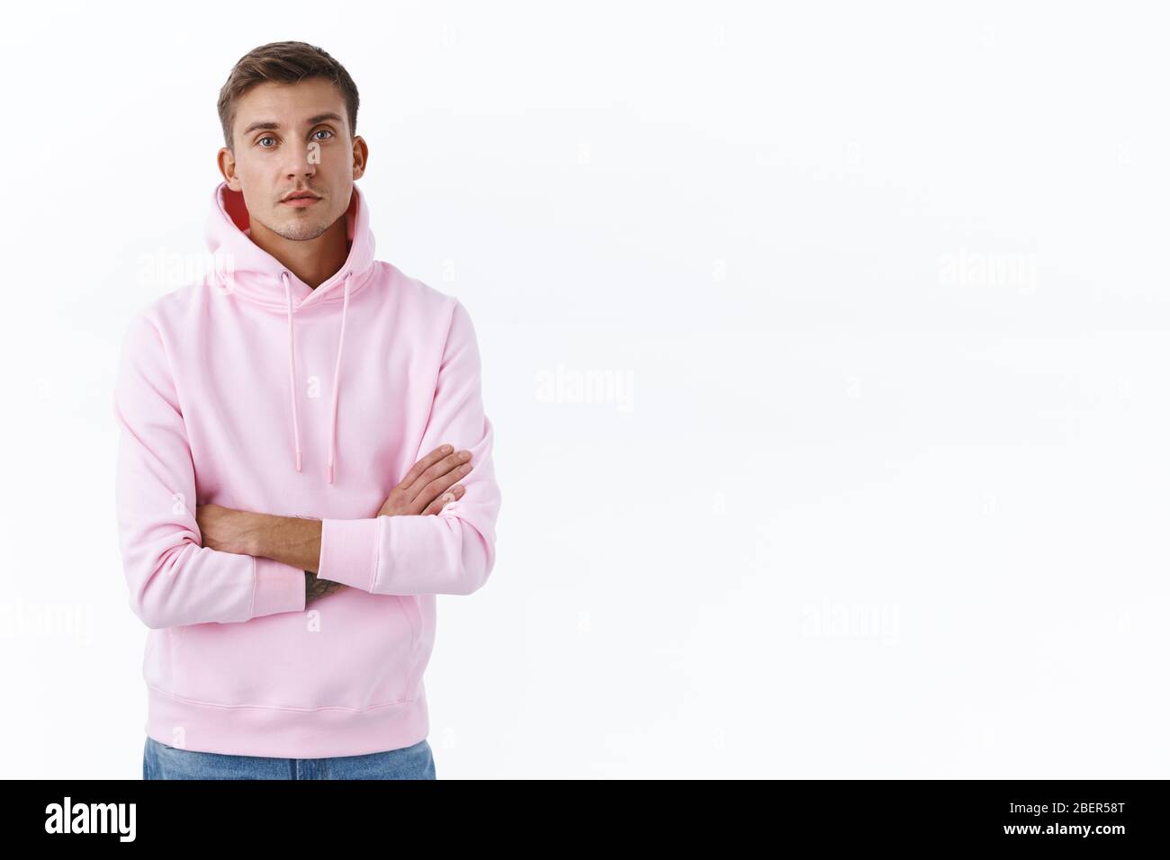 Ritratto di uomo biondo spensierato e rilassato con felpa con cappuccio rosa, braccio trasversale sopra il petto e guardare fotocamera seria, ottenendo diritto al lavoro Foto Stock