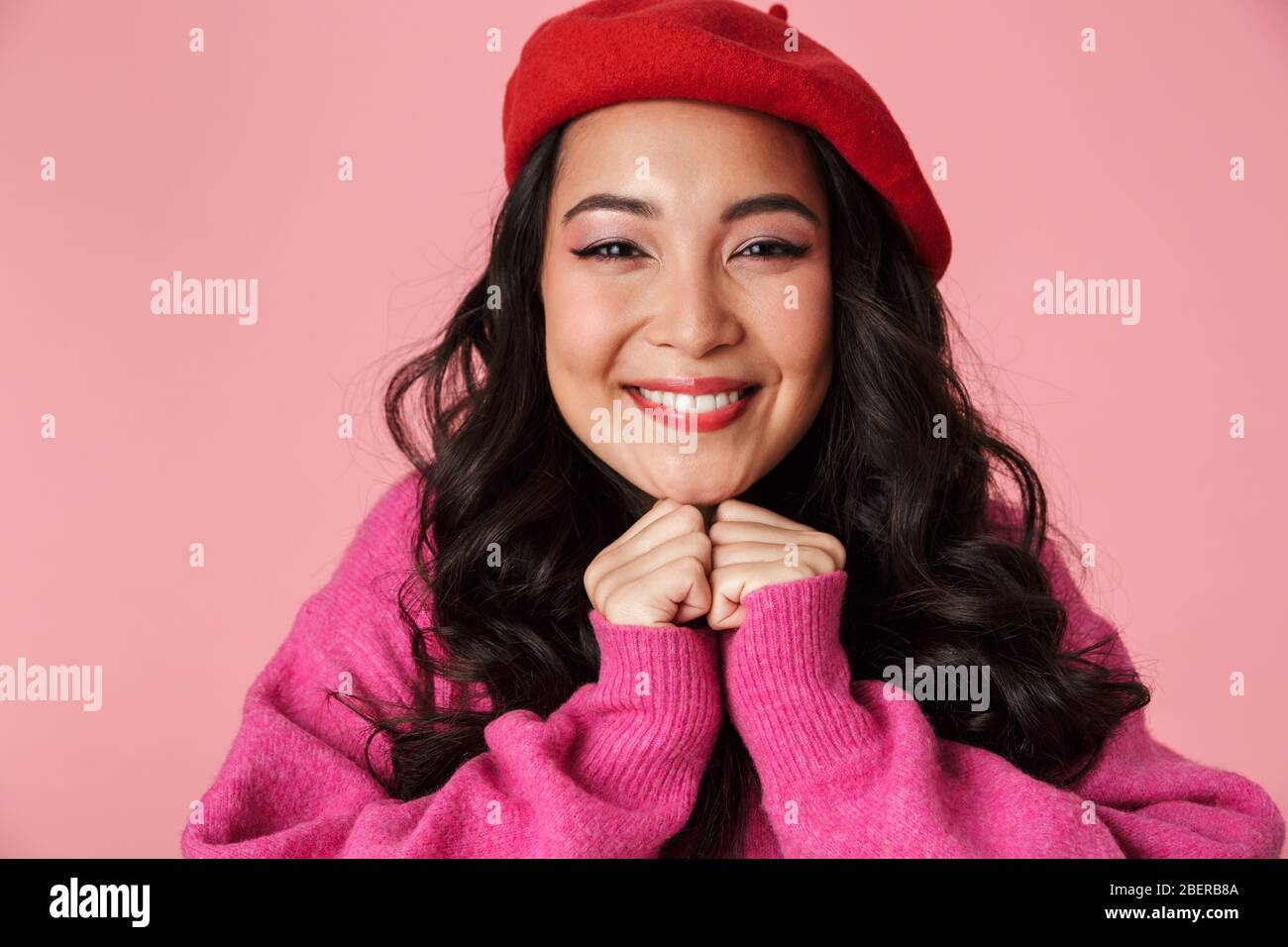 Immagine di giovane bella ragazza asiatica che indossa beret sorridente e clenching le sue pugni al petto isolato su sfondo rosa Foto Stock