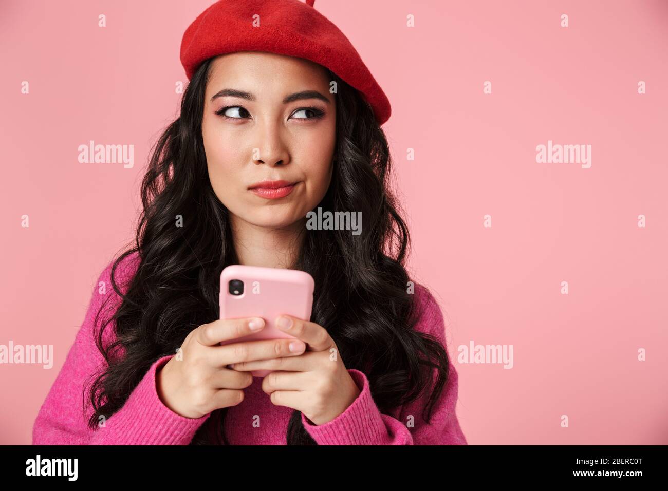 Immagine di una bellissima ragazza asiatica perplessa che indossa beret accigliato e tenendo smartphone isolato su sfondo rosa Foto Stock