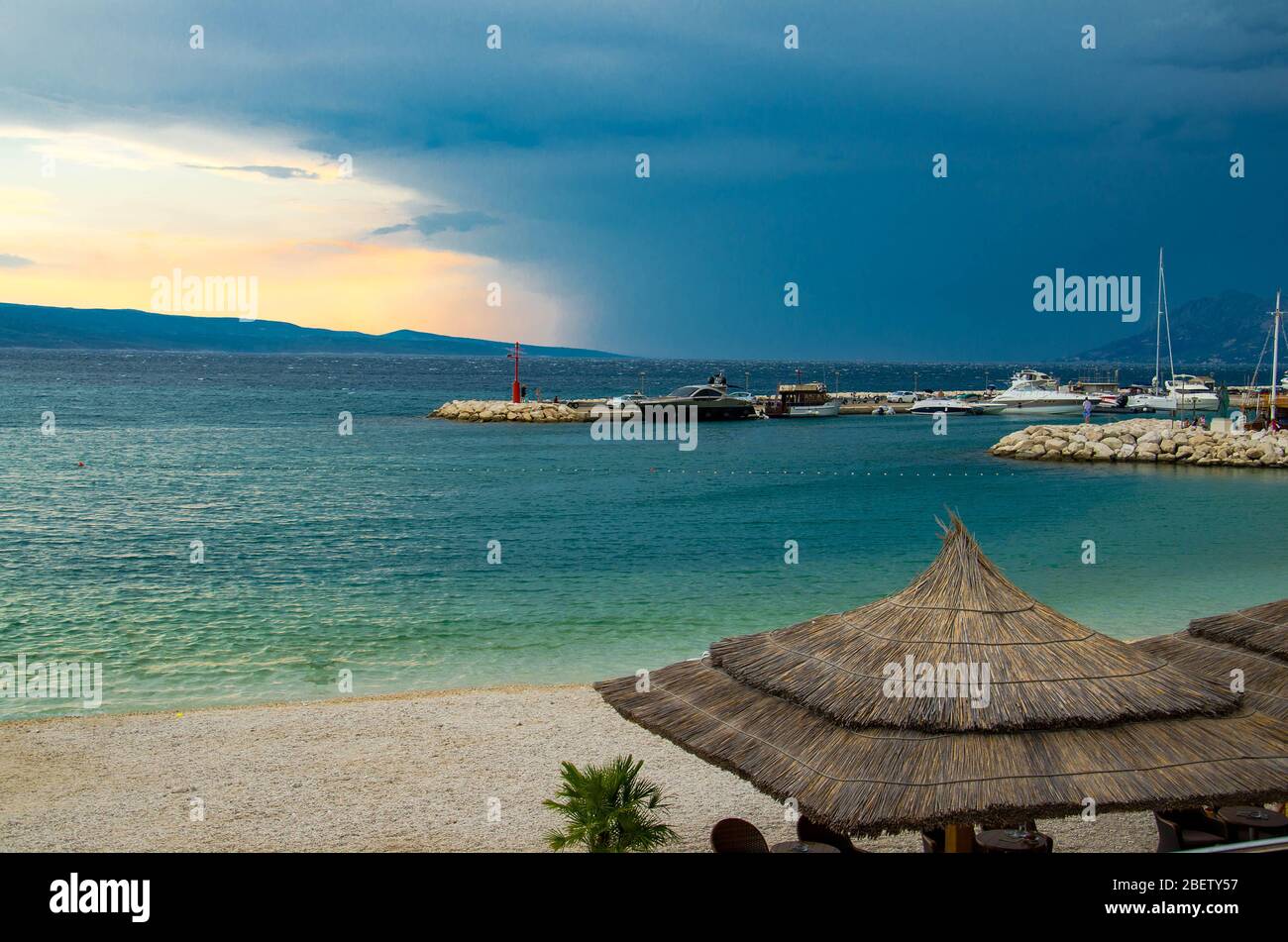 Bella vista della spiaggia di sabbia con ombrelloni paglia, porto e piccolo faro sul molo in pietra di fronte all'isola di Brac alla vigilia della pioggia, Baska Vo Foto Stock