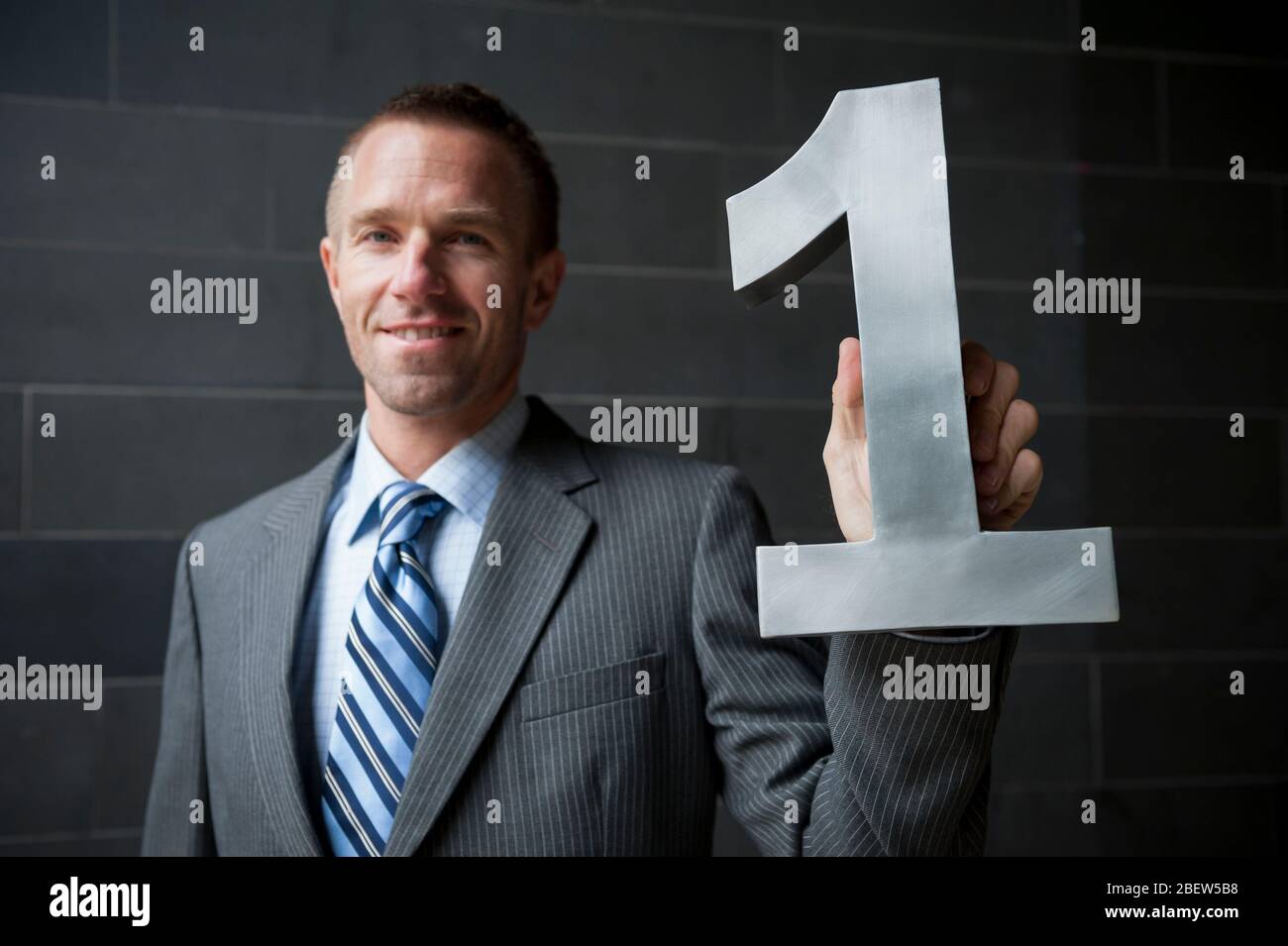 Uomo d'affari sorridente in una tuta che tiene un grande numero di acciaio uno con sfondo scuro Foto Stock