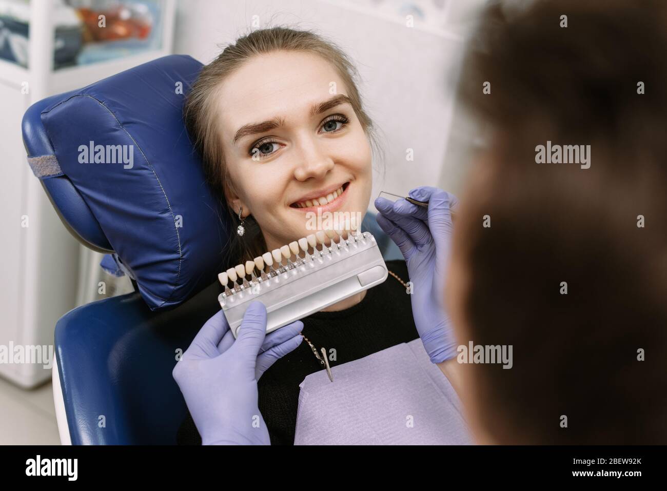 Dentista in guanti medici blu applicando un campione dalla scala di smalto dei denti a denti di una paziente felice per raccogliere l'ombra destra, procedura di sbiancamento dei denti, Foto Stock