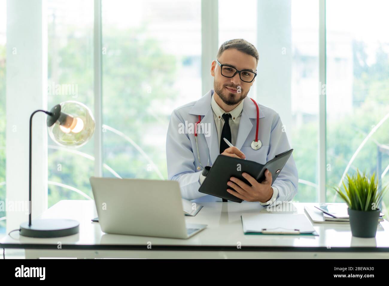 Dottore seduto alla scrivania al suo posto di lavoro e sorridente alla macchina fotografica. Servizio medico perfetto in clinica. Felice futuro della medicina e della sanità. Foto Stock
