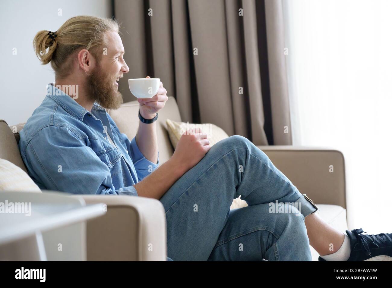 Un giovane uomo felice e rilassato lounge sul comodo divano che beve il tè. Foto Stock