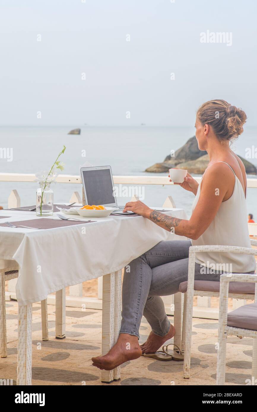 Una donna che beve un caffè con il suo computer portatile seduto in un ristorante vicino alla spiaggia una mattina presto. Foto Stock