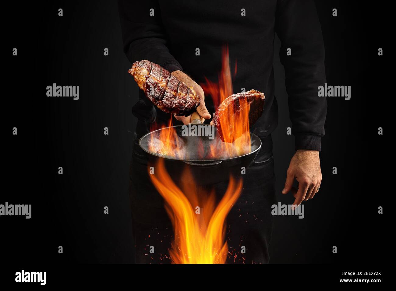 Giovane maschio vestito con jumper e jeans. Sta tenendo la pentola del wok bruciante con il fumo sopra il fuoco e friggere due bistecche di manzo, sfondo nero. Primo piano Foto Stock