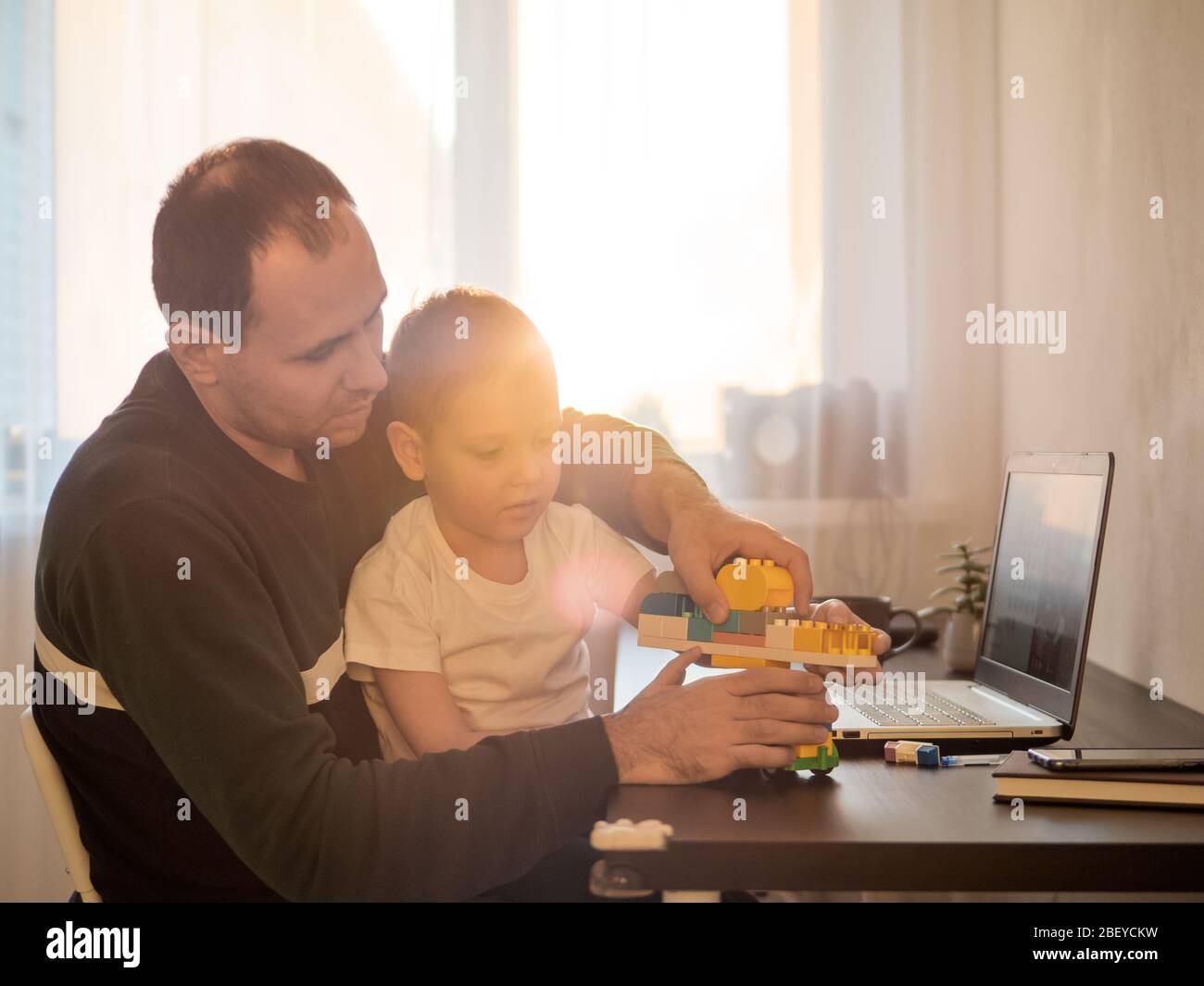 Quattro anni ragazzo giocare e padre al tavolo con notebook. Lavoro a domicilio durante il periodo di isolamento della quarantena del coronavirus. Lavorare tra i bambini, lavorare a distanza concetto. Luce del tramonto Foto Stock