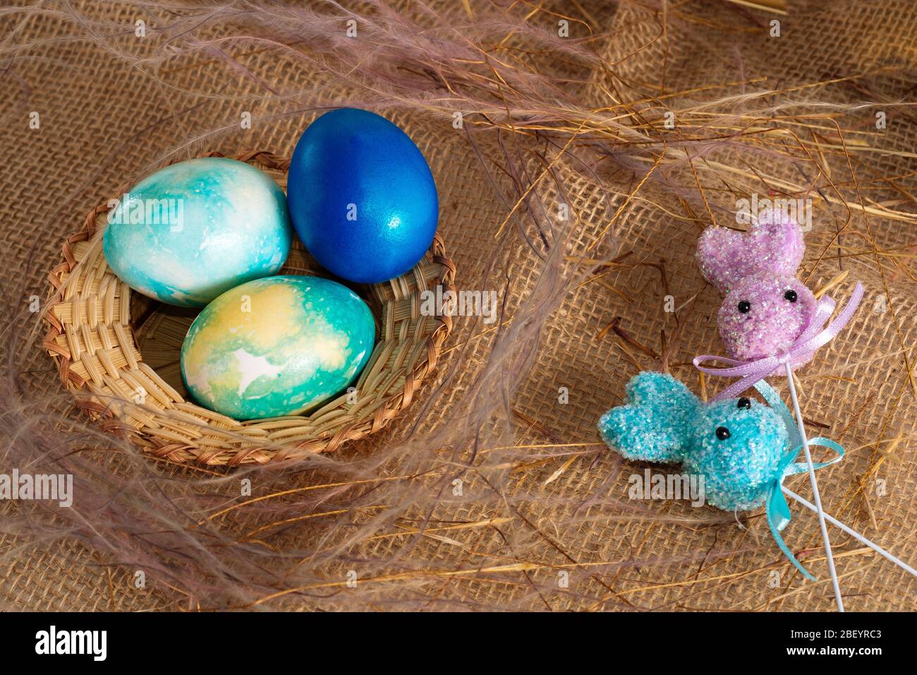 Uova pasquali multicolore dai toni blu e due coniglietti pasquali su un bastone su uno sfondo di stoffa marrone. Foto Stock