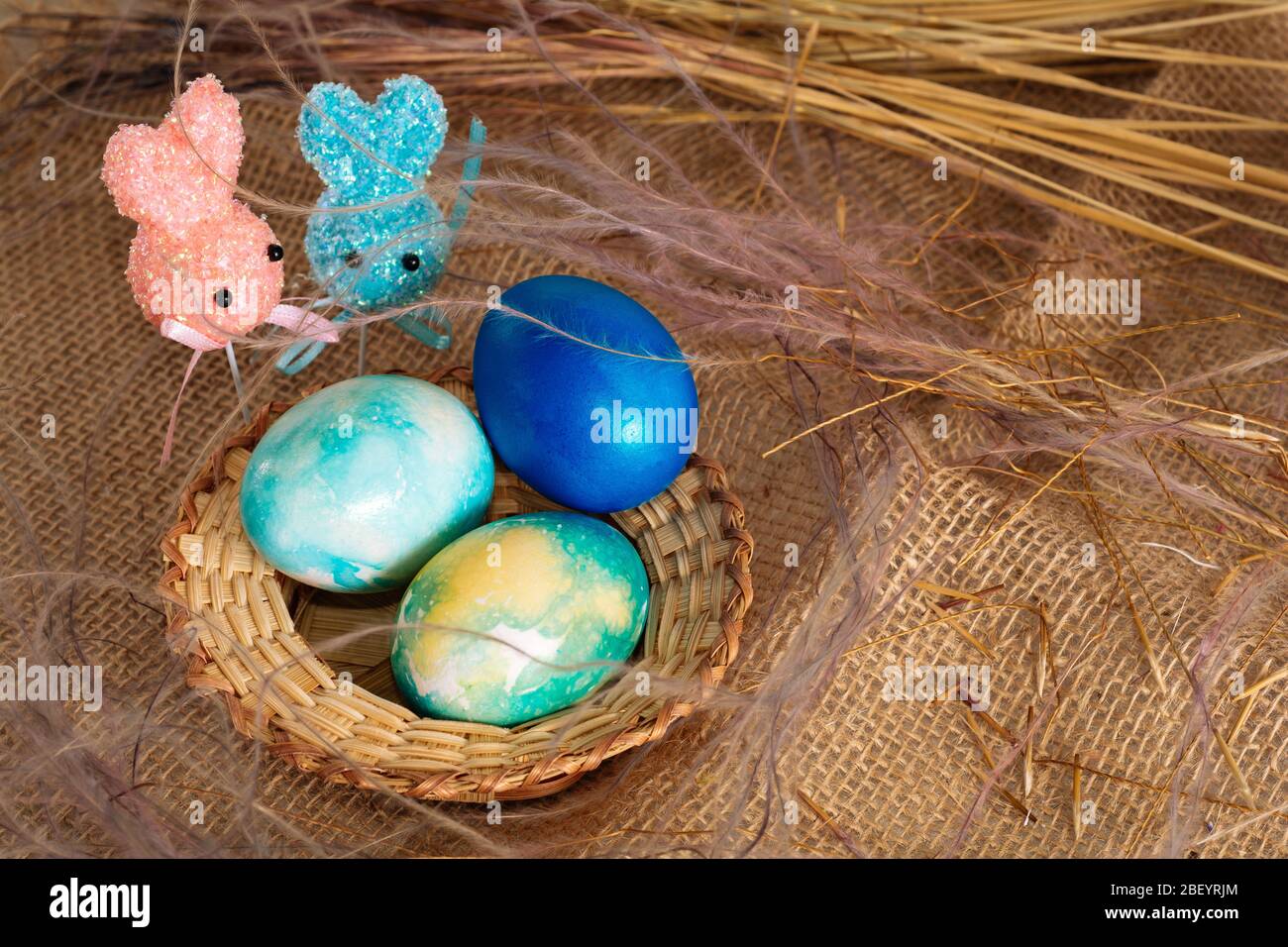 Uova pasquali multicolore dai toni blu e due coniglietti pasquali su un bastone su uno sfondo di stoffa marrone. Foto Stock