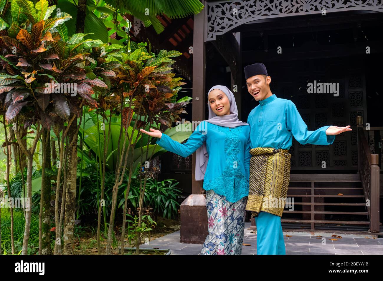 Un ritratto di giovane coppia di malay muslim in costume tradizionale durante la celebrazione di Aidilfitri che mostra un gesto di benvenuto di legno tradizionale Foto Stock
