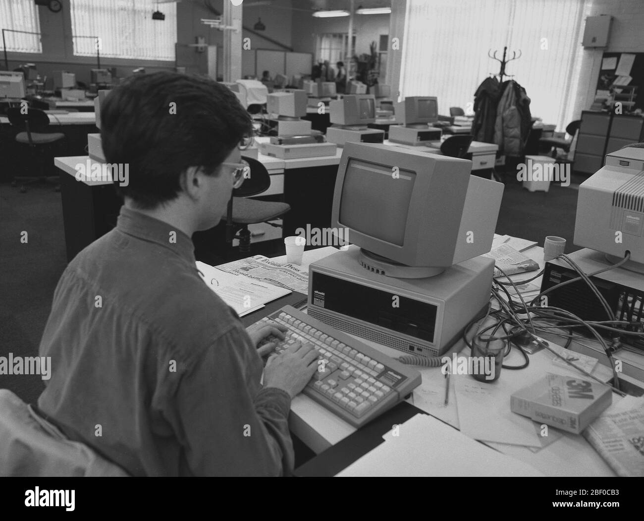 1987, un giornalista maschio che stenta a una scrivania usando un personal computer, noto come PC, in un ufficio. I PC sono visibili sulla maggior parte delle scrivanie. I giornali e l'industria della stampa sono stati tra i primi a realizzare i potenziali vantaggi e il risparmio sui costi derivanti dall'utilizzo dei nuovi personal computer, consentendo di produrre copie in modo più efficiente ed economico rispetto al metodo tradizionale ad alta intensità di lavoro della composizione dei metalli. Foto Stock