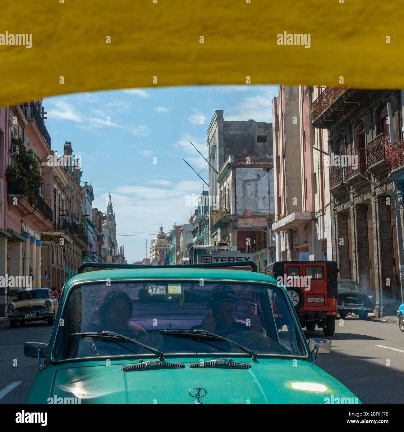 Coppia in una lada auto dando i pollici in su ai turisti che cavalcano nella parte posteriore di un cocotaxi giallo attraverso la città vecchia di Havana, Cuba Foto Stock