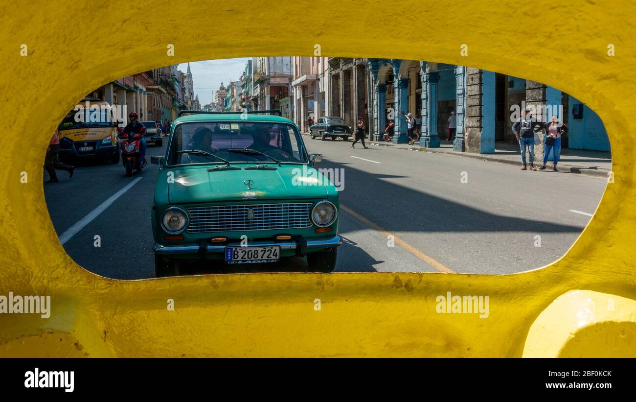Vista di una coppia in un'auto lada che guida dietro un cocotaxi giallo attraverso la città vecchia di Havana, Cuba Foto Stock