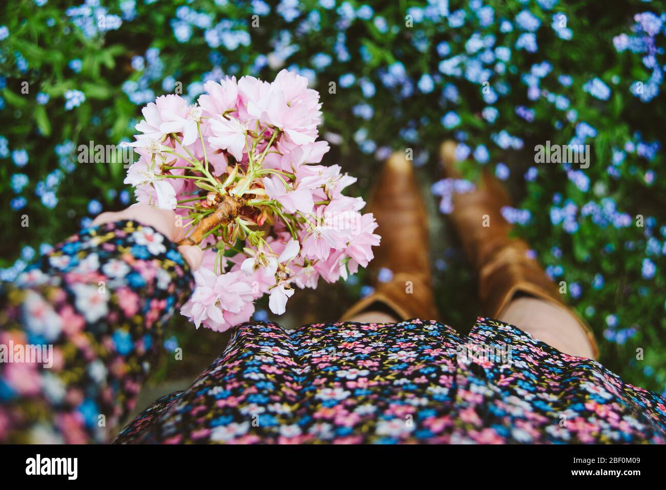 Una donna o una ragazza che si trova all'esterno in una giornata primaverile indossando un abito floreale e stivali cowboy e tenendo rosa fioritura, fresco e floreale. Dof poco profondo. Foto Stock