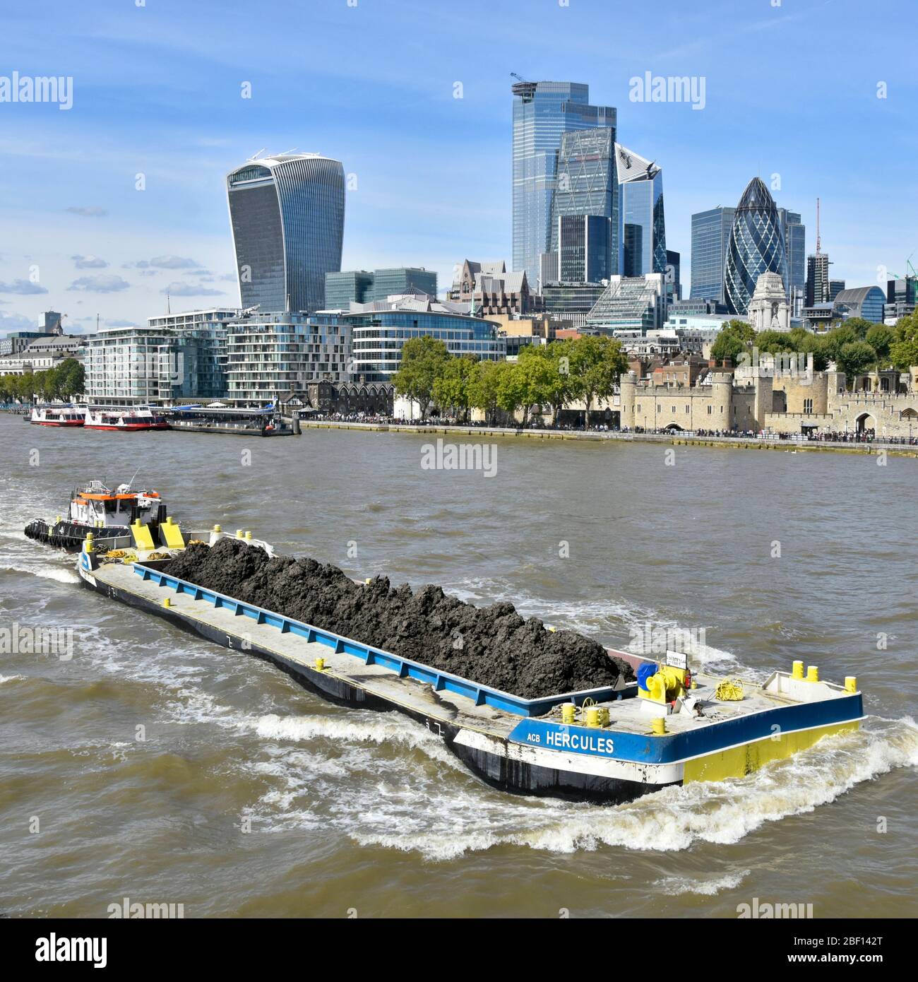 Trasporto fluviale lunga chiatta e carico di terreno scavato dal progetto di fognatura Thames Tideway Tunnel spinto da una barca di rimorchiatore che passa City of London skyline Inghilterra UK Foto Stock