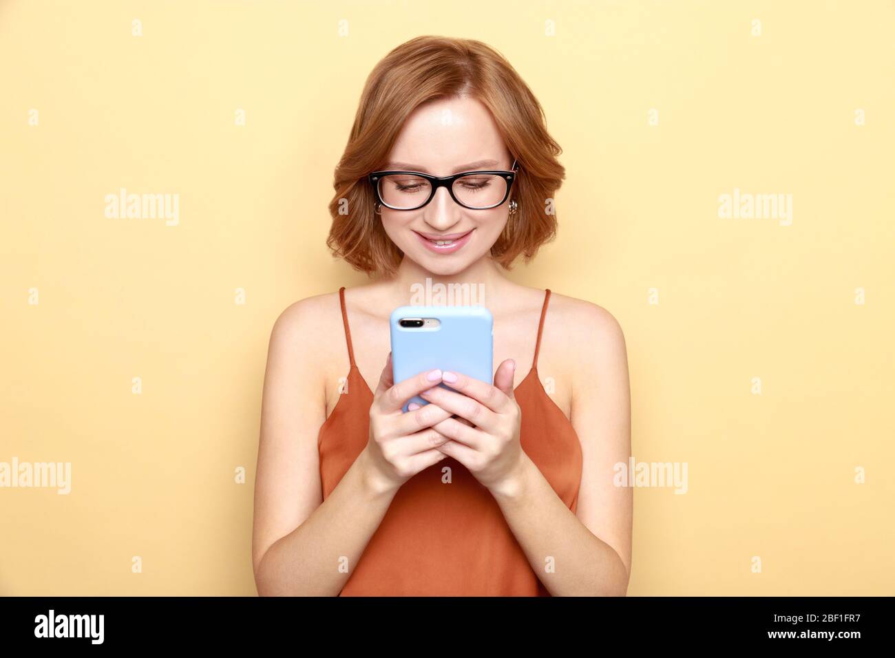 Studio ritratto di sorridente bella giovane donna che indossa occhiali, guarda e scrive un messaggio sul suo smartphone al suo ragazzo o controllare i social media, isolat Foto Stock